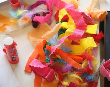 Kolorowe paski bibuły i piórka rozsypane na stole, obok klej w sztyfcie. Materiały do prac plastycznych.