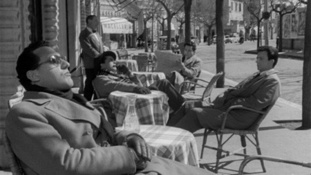 [Czarno-biały kadr z filmu. Czterech mężczyzn siedzi przy stolikach restauracyjnych.]