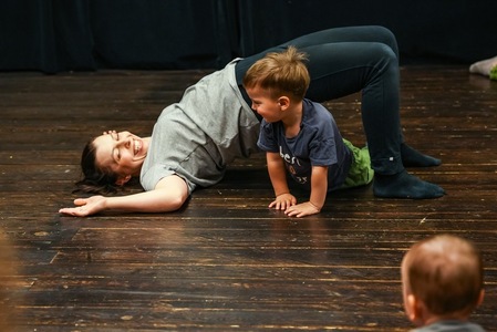 Uśmiechnięta kobieta bawi się na podłodze z małym chłopcem. Chłopiec przechodzi pod jej uniesionymi nogami.