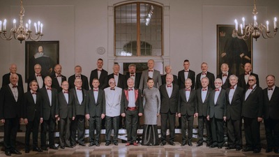 Chór Męski Copernicus - jego członkowie stojący w czarnych garniturach. Stoją w dwóch szeregach w zabytkowej sali jakiegoś pałacu. W tle duże portrety właścicieli pałacu.