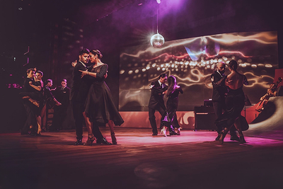 Na scenie tancerze w eleganckich, czarnych strojach wykonują tango. Kobiety ubrane w dopasowane suknie. Mężczyźni w czarnych garniturach prowadzą zdecydowanym, eleganckim gestem.