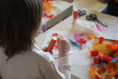 Dziecko siedzi przy stoliku i tworzy pracę plastyczną, używając kleju w sztyfcie. Na blacie leżą kolorowe piórka, kawałki bibuły, nożyczki i inne materiały do zabawy twórczej.