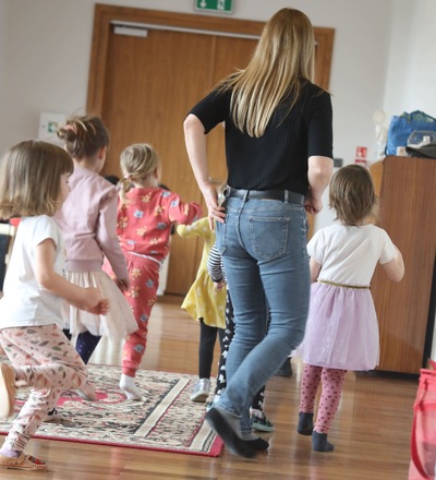 Dzieci poruszają się po sali razem z dorosłą osobą, która prowadzi zajęcia. Idą jedno za drugim po dywanie i podłodze. Widać drzwi i jasne wnętrze sali.