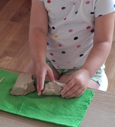 Dziewczynka siedzi przy stole i lepi z gliny na zielonej podkładce. W dłoniach trzyma kawałek jasnej masy, obok leżą inne fragmenty. Ubrana w koszulkę w kolorowe kropki.