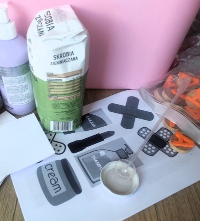 Na blacie stoi paczka skrobi ziemniaczanej, butelka kosmetyku i plastikowy pojemnik. Na dole leży zakrętka z małą miarką oraz pipetą, a obok znajduje się woreczek z drewnianymi, pomarańczowymi literkami. Zdjęcie zrobione z góry.