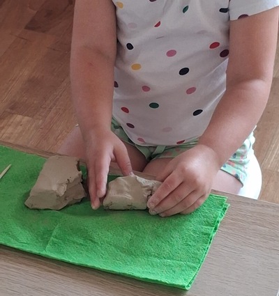 Dziewczynka siedzi przy stole i lepi z gliny na zielonej podkładce. W dłoniach trzyma kawałek jasnej masy - gliny, obok leżą inne fragmenty gliny. Ubrana w koszulkę w kolorowe kropki.