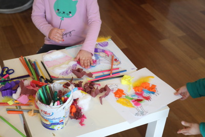 Dziecko siedzi przy stole i robi pracę plastyczną. Na stole leżą kredki, flamastry, nożyczki i kolorowe kawałki papieru. Widać rysunek oraz watę przyklejoną do kartki.