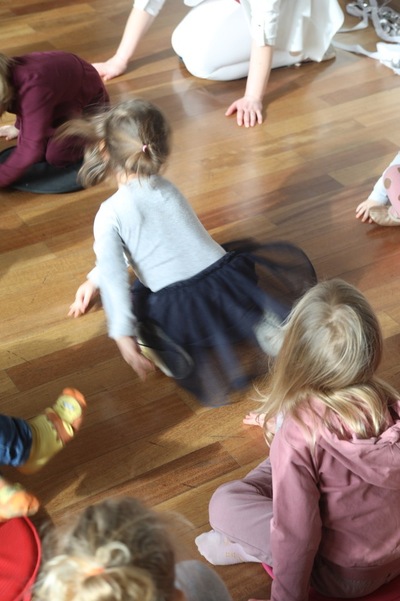 Dzieci w ruchu na drewnianej podłodze podczas zajęć ruchowych; uczestnicy siedzą i poruszają się w kręgu, w tle osoba prowadząca.