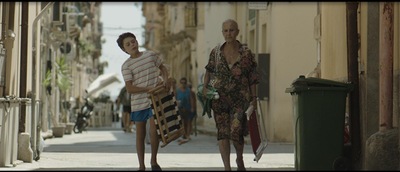[Kolorowy kadr z filmu. Chłopiec idzie z siwą kobietą ulicą.]