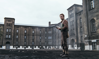 Kolorowy kadr z filmu. Mężczyzna w eleganckim ubraniu, z książką w dłoni, stoi na tle monumentalnego budynku z cegły.