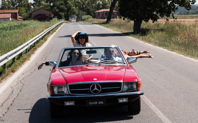 Kolorowy kadr z filmu. Cztery osoby, trzy kobiety i mężczyzna, jadą czerwonym kabrioletem pustą ulicą.