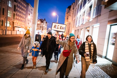 Grupa dzieci i dorosłych. Jedna osoba ma podniesioną rękę, w której trzyma tabliczkę.
