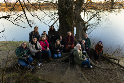 Na środku znajduje się grupa osób z aparatami fotograficznymi. W tle drzewo przy konarach, którego siedzą a za nim rzeka.