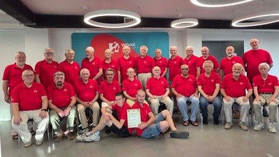 Na zdjęciu widać chór składający się z 25 mężczyzn, pozujących do zdjęcia w korytarzu budynku Młyn Kultury. Stoją w dwóch rzędach. Ubrani są w czerwone koszulki typu polo z małym logiem Copernicus na lewej kieszonce.