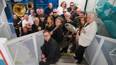 Grupa muzyków w różnym wieku. Są to zarówno kobiety i mężczyźni.Maja oni na sobie czarne i białe stroje. Kilka osób trzyma w rękach trąbki, saksofony i klarnety. Cala grupa stoi na schodach na półpiętrze. W tle za szybami panorama miasta