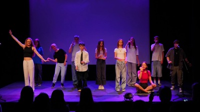 Grupa młodych dziewczyn i chłopaków w czasie przedstawienia teatralnego. Dziewczyna po lewej stronie ma prawą rękę skierowaną do góry. Po prawej stronie jedna z dziewczyn siedzi na po turecku. Dwóch chłopaków za nią mówi przez mikrofony.