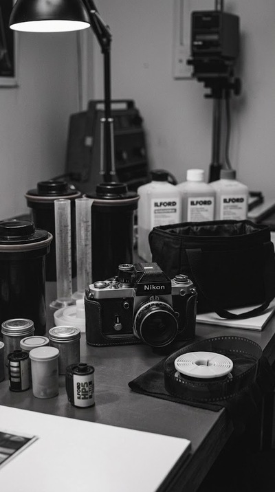 Czarno-białe zdjęcie przedstawiające pracownię do wywoływania filmów analogowych. Na pierwszym planie stoi klasyczny aparat Nikon w skórzanym etui, otoczony akcesoriami, w tym szpulą z nawiniętym negatywem.