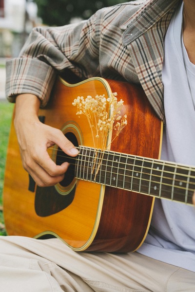 Zbliżenie na osobę, która trzyma na kolanach drewnianą gitarę akustyczną. Prawa dłoń spoczywa na strunach tuż nad otworem rezonansowym. W struny wpleciona jest delikatna gałązka drobnych, jasnych, suszonych kwiatów.