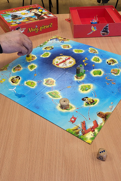 Rozgrywka w grę planszową. Plansza imituje ocean z wieloma ponumerowanymi polami, po których poruszają się miniaturowe plastikowe pionki. Dłoń w lewym górnym rogu przesuwa żółty pionek. Na drewnianym stole leży drewniana kostka do gry.