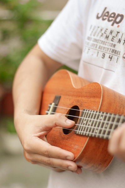 Zbliżenie na osobę w białej koszulce, trzymającą drewniane ukulele. Widoczna dłoń opiera się na pudle rezonansowym w pobliżu strun, delikatnie przytrzymując instrument blisko tułowia.