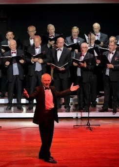 Chór Męski Arion na scenie w zamkowej sali, podczas koncertu, pod batutą dyrygenta. Dyrygent stoi zwrócony do publiczności.