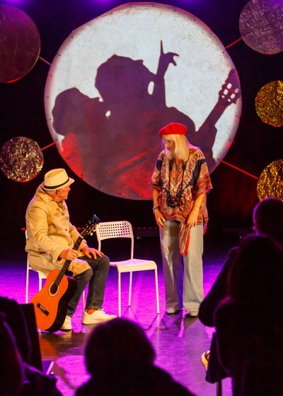 Dwie osoby występujące na scenie podczas zajęć teatralnych, jedna siedzi z gitarą na krześle. W tle duża okrągła dekoracja świetlna symbolizująca układ planet.